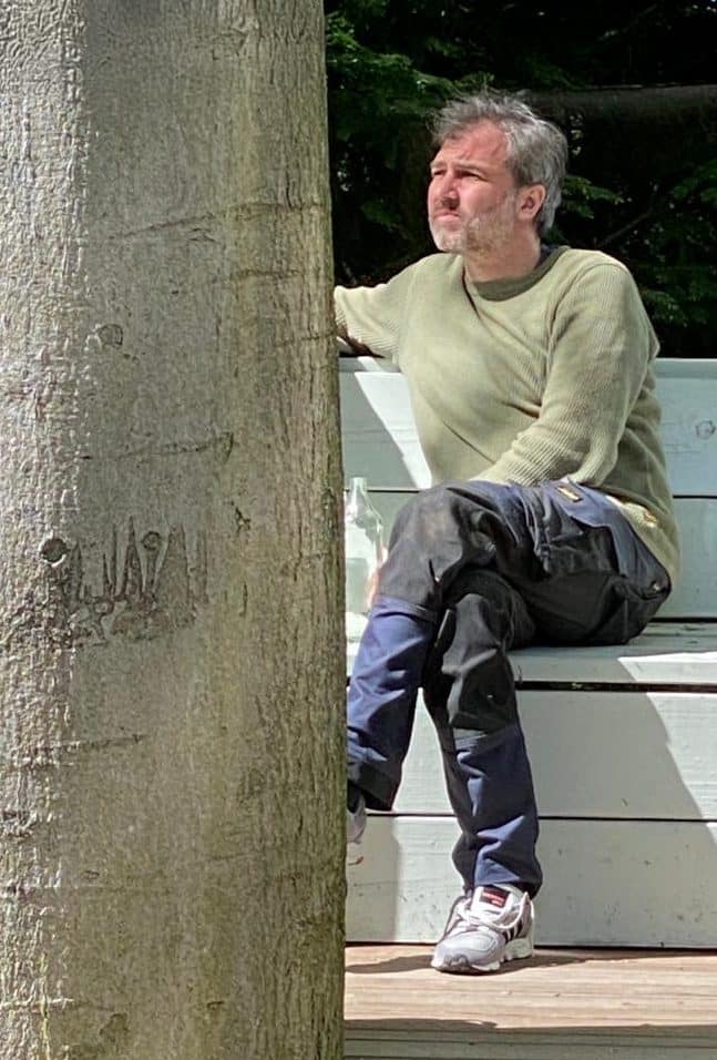 Ein Mann sitzt seitlich auf einer weißen Bank, der Blick wird durch einen dicken Baumstamm im Vordergrund teilweise verdeckt.