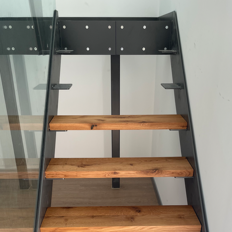 Eine offene Holztreppe mit schwarzem Stahlrahmen und Glasgeländer steht vor einer hellen Wand.