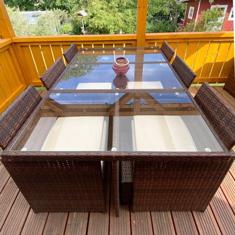 Ein Gartentisch aus Rattan trägt eine klare Glasplatte unter freiem Himmel.