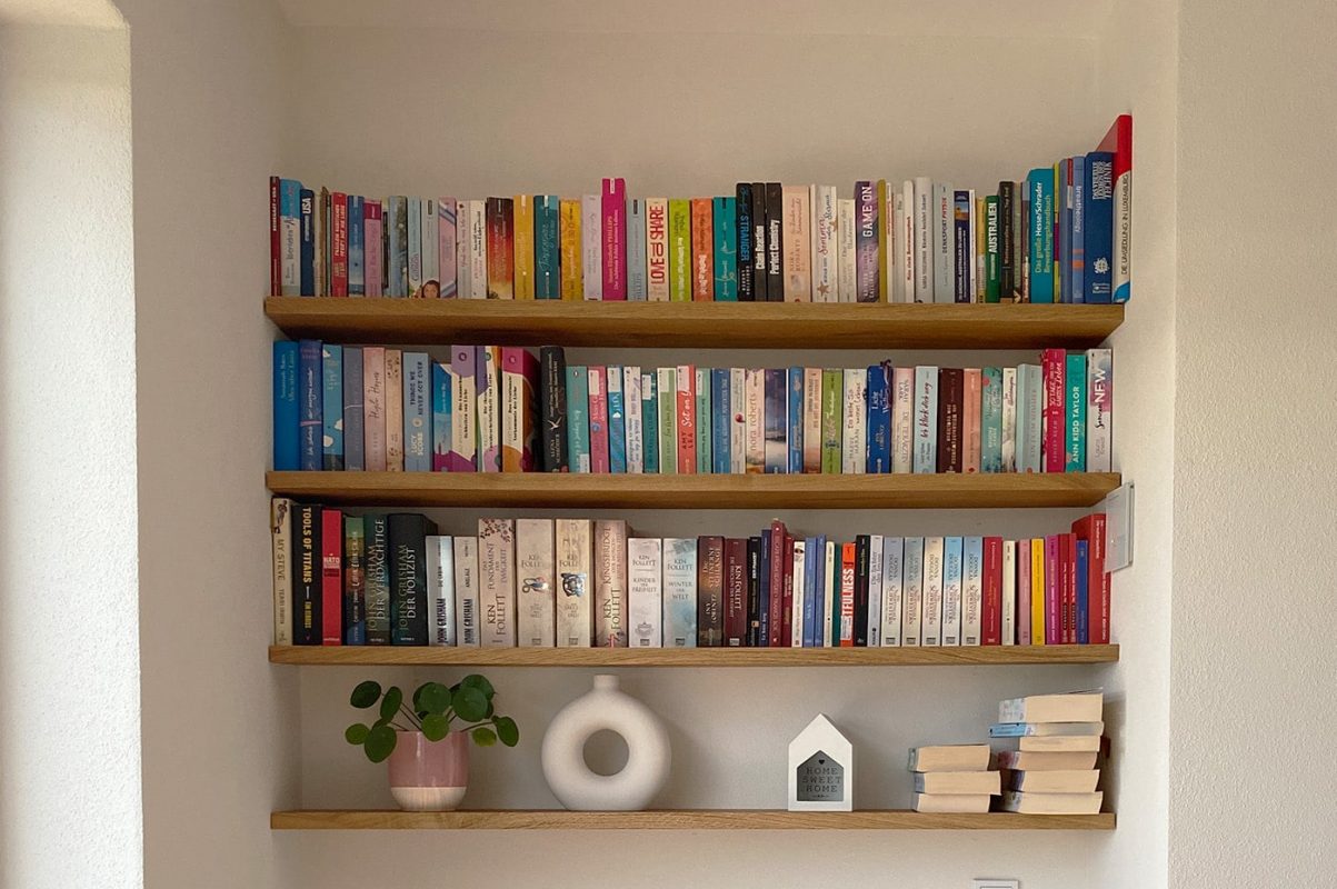Ein Wandregal trägt zahlreiche Bücher und dekorative Objekte in einem Wohnraum.