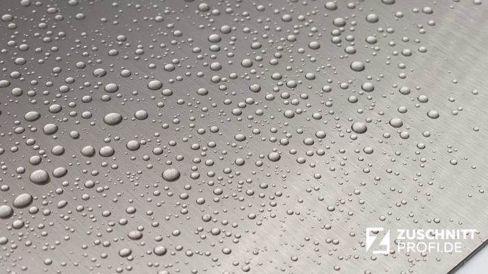 Wasserperlen auf gebürsteter Edelstahloberfläche mit dem weißen zuschnittprofi.mom-Logo auf der unteren rechten Seite.