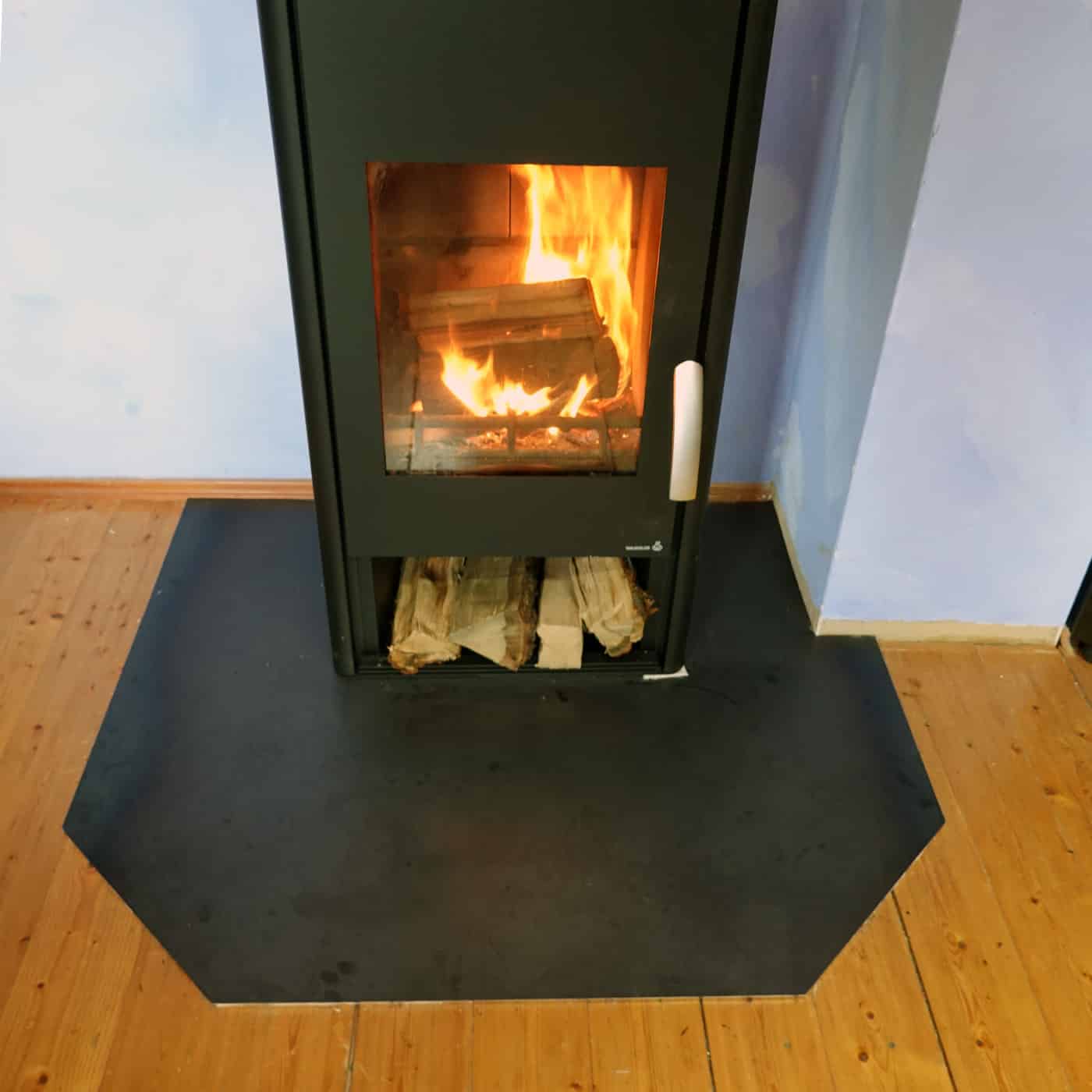 Eine kantige Funkenschutzplatte liegt vor einem brennenden Kamin auf hellem Holzboden.
