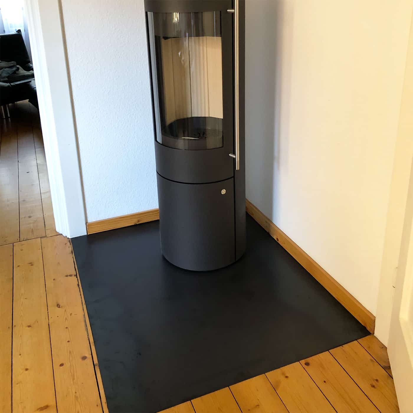 Eine schwarze Funkenschutzplatte liegt passgenau in einer Raumecke auf dem Holzboden.