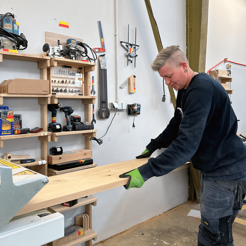 Eine Handwerkerin bearbeitet eine Holzplatte in einer aufgeräumte Werkstatt.