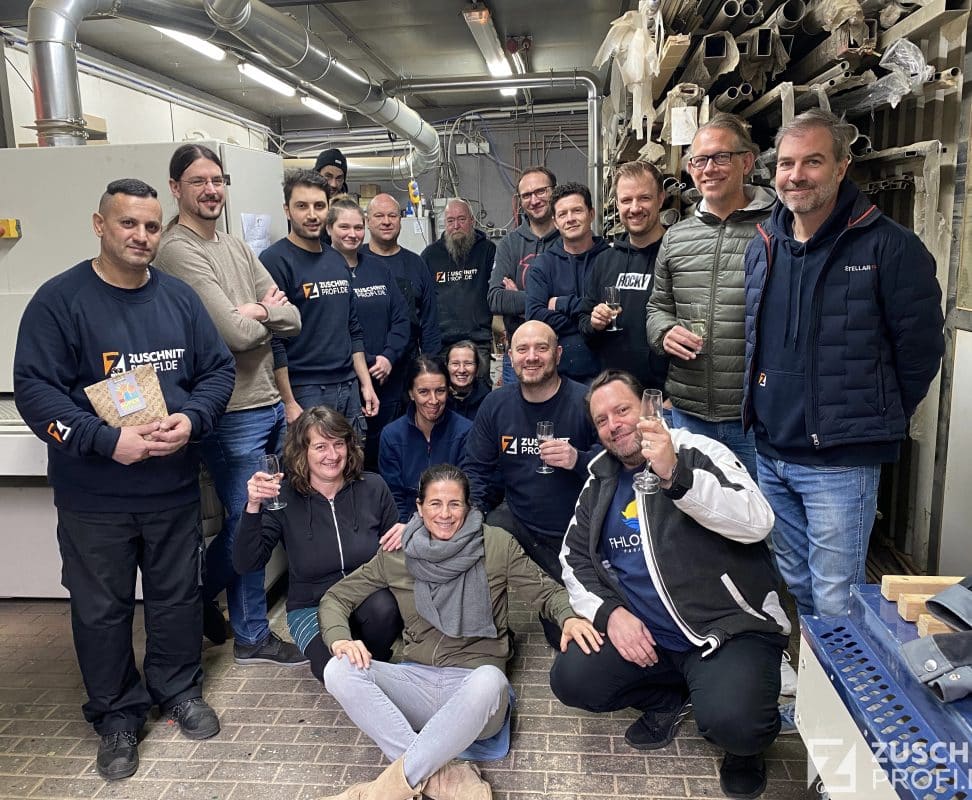 Eine größere Gruppe von Zuschnittprofi-Mitarbeitern steht lächelnd zusammen in einer metallverarbeitenden Werkstatt.