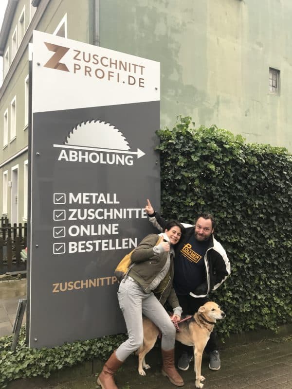 Zwei Personen und ein Hund posieren vor dem Zuschnittprofi-Abholschild.