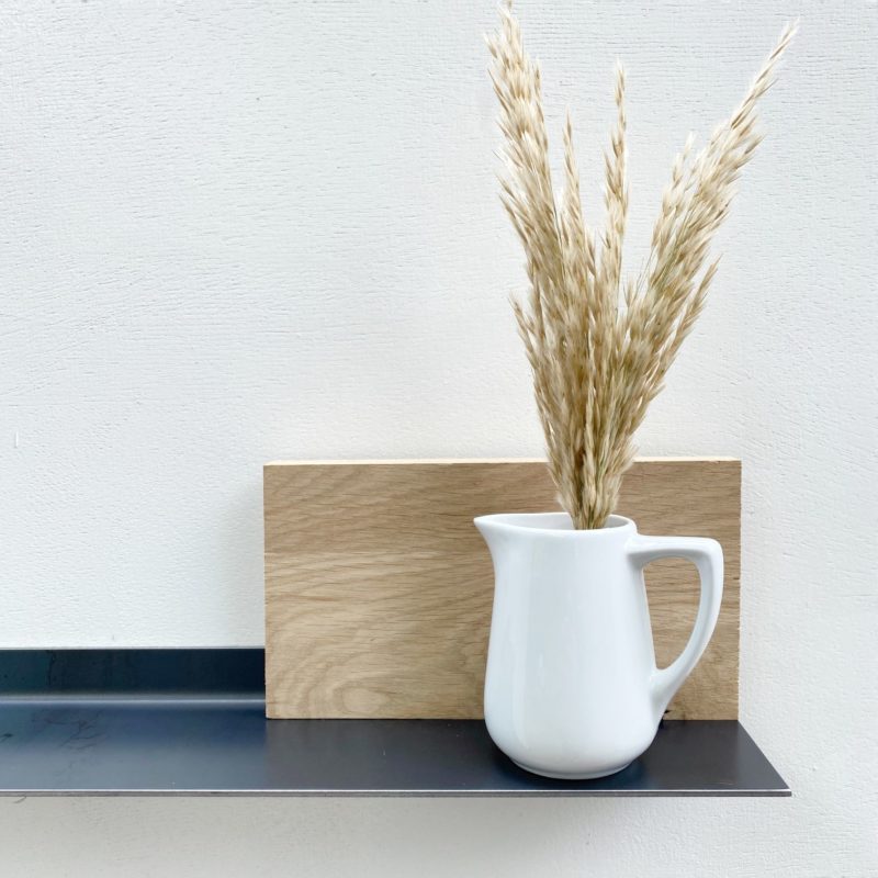 Weiße Vase mit Trockenblumen vor Holzplatte auf schwarzem Regal.