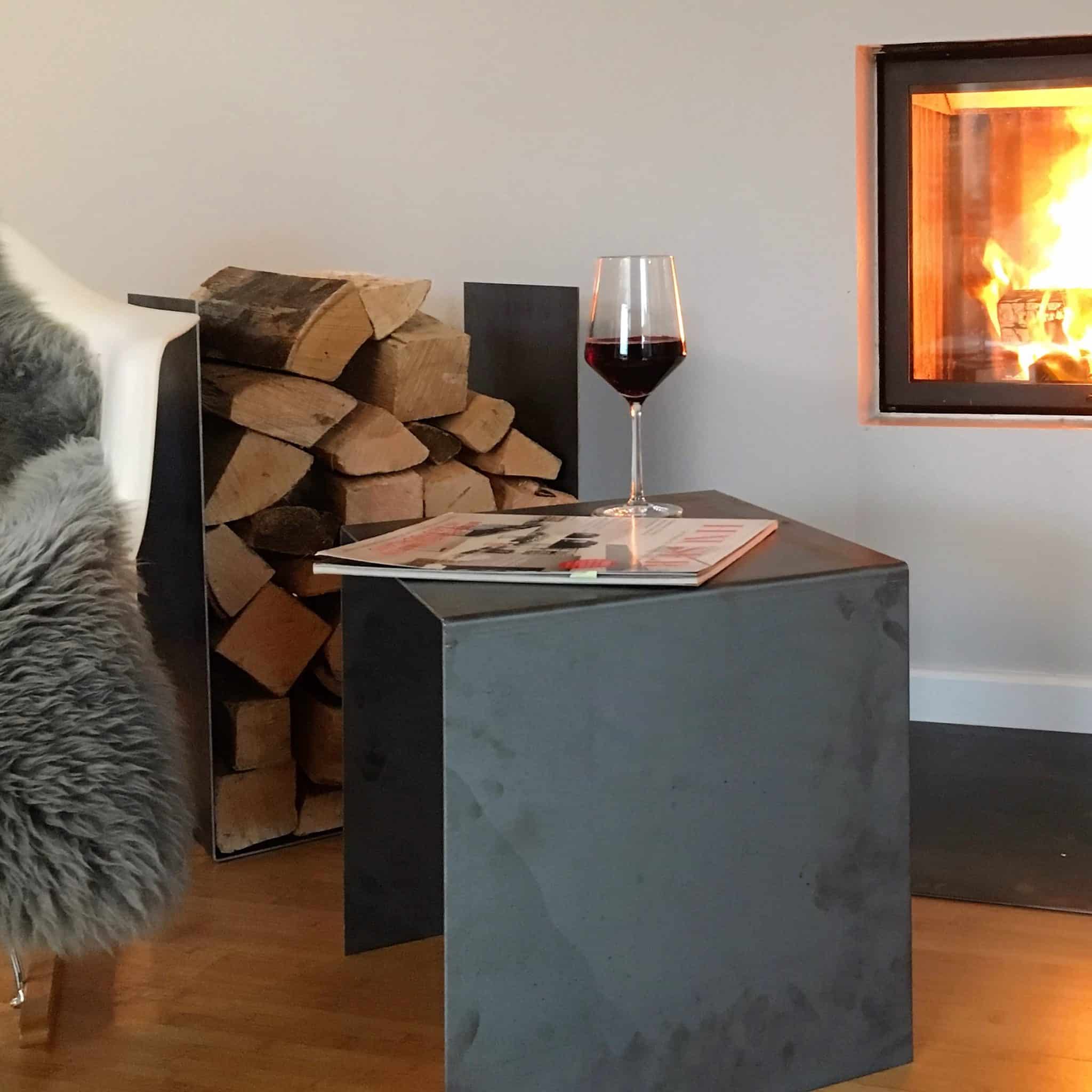 Ein kleiner Beistelltisch aus dunkelgrauem Stahl steht neben einem Kamin, daneben liegt gestapeltes Kaminholz.