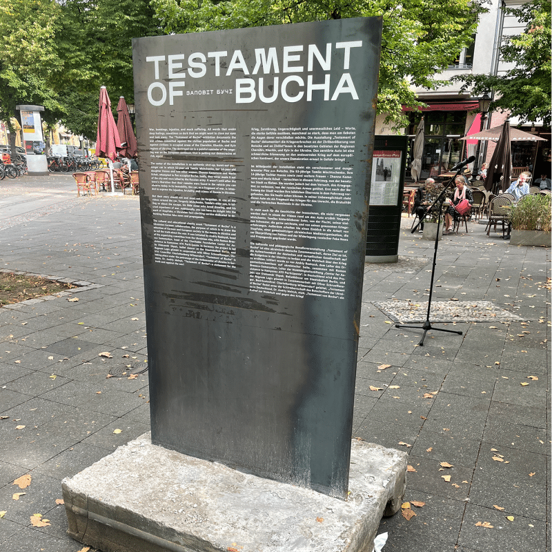 Eine beschriftete Metallstele steht auf einem Platz mit Außengastronomie umgeben.