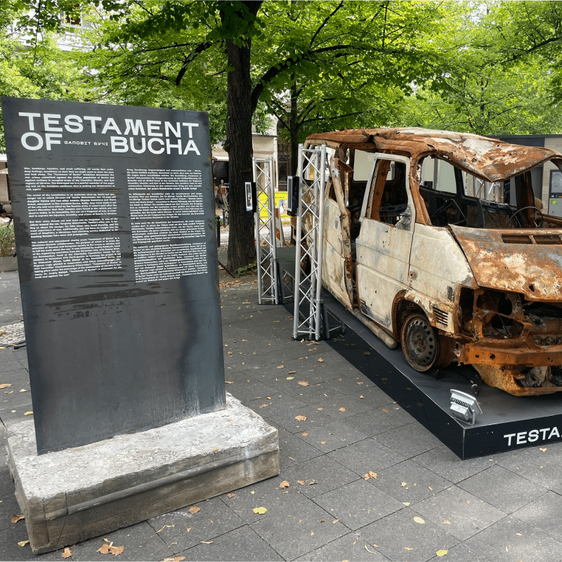 Eine beschriftete Metallstele steht draußen vor einem nicht funktionstüchtigen Transporter auf einem Platz.