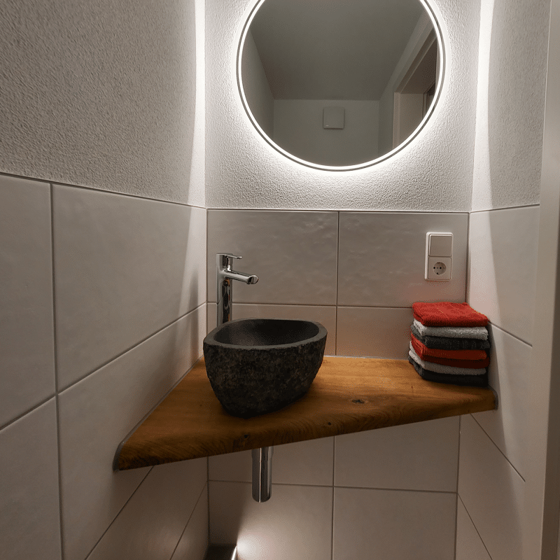 Eine kleine Holzablage mit integriertem Waschbecken ist unter einem runden Spiegel im Bad montiert.