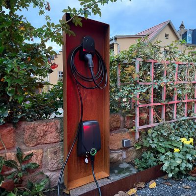 Eine freistehende Cortenstahlstele trägt eine Wallbox und setzt einen markanten Akzent im Garten.