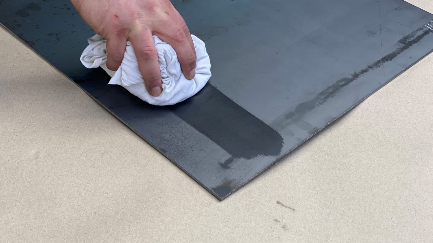 Eine Hand ölt eine schwarze Funkenschutzplatte auf hellem Untergrund.