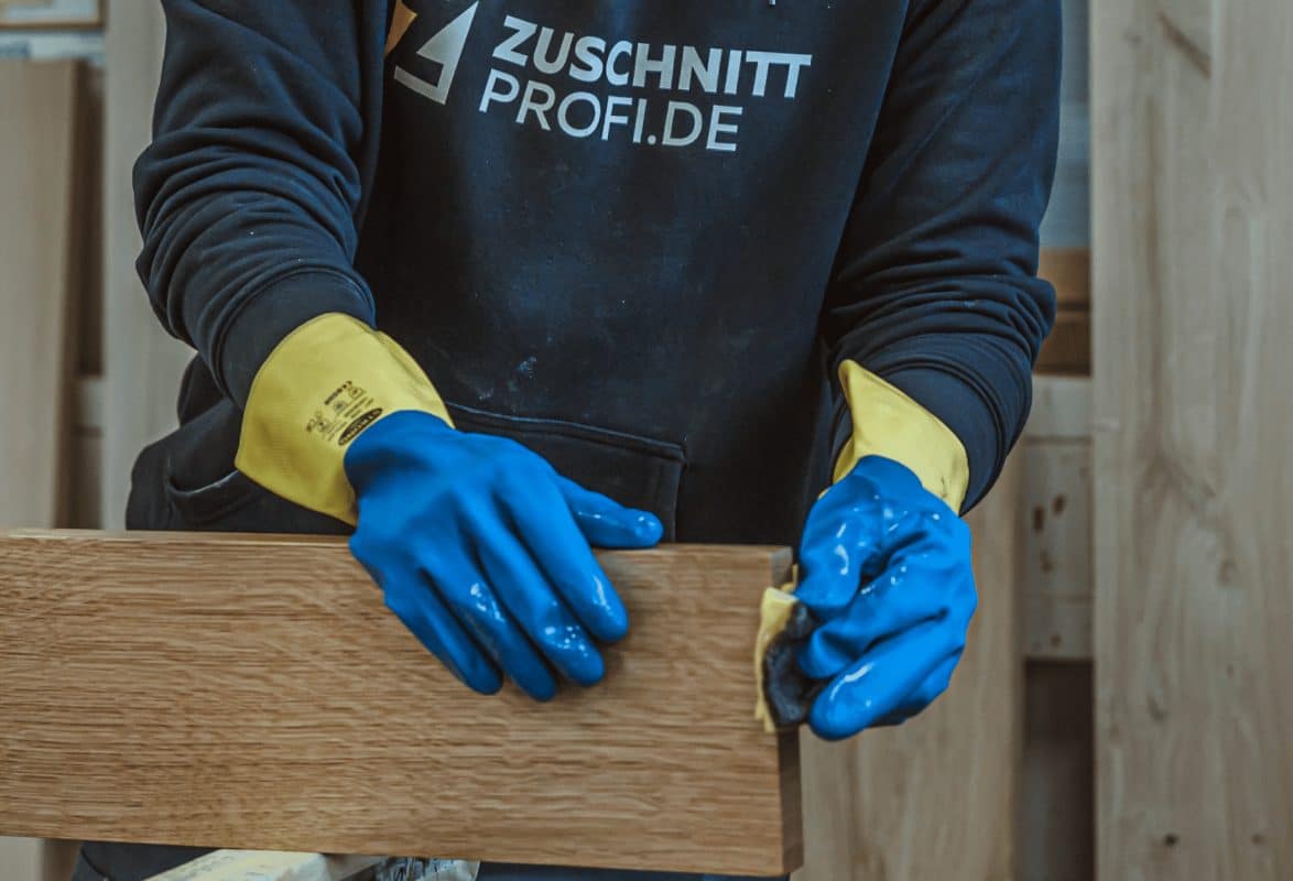 Ein Mitarbeiter von zuschnittprofi.mom arbeitet mit Handschuhen an einem Holzstück und verarbeitet die Oberfläche mit ruhiger Hand.