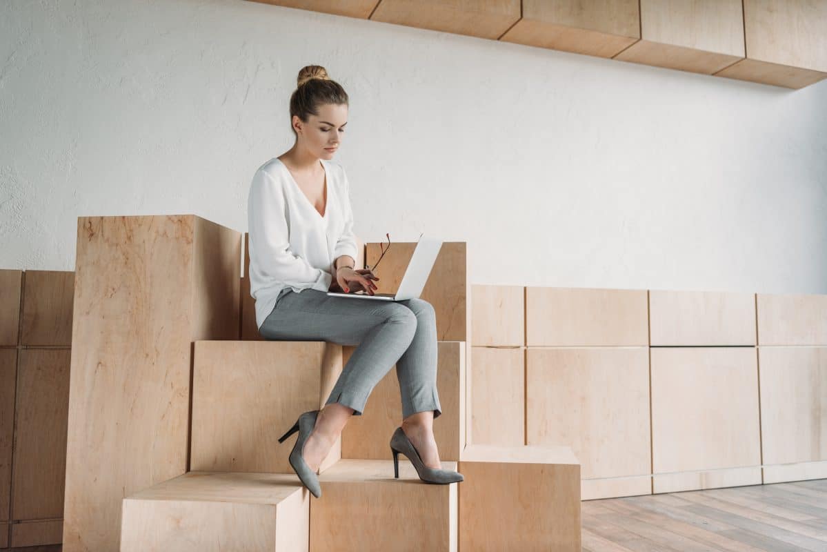 Frau sitzt mit Laptop auf minimalistischer Sitzbox aus hellem Holz.
