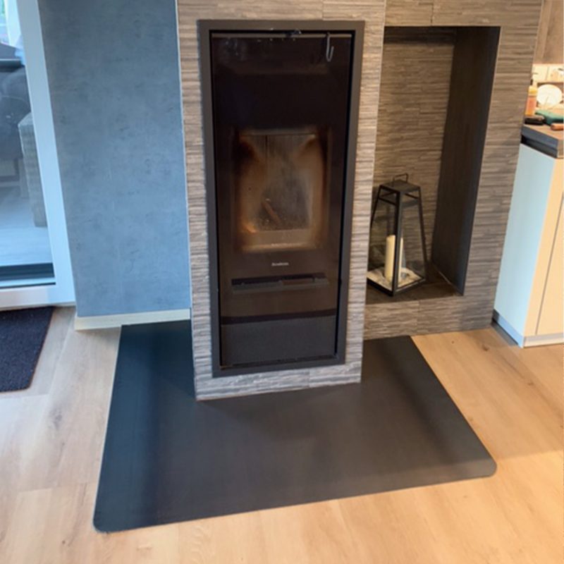 Abgerundete Funkenschutzplatte aus Schwarzstahl mit Ausschnitt, platziert auf dem Boden vor einem Kamin als Funkenschutz.