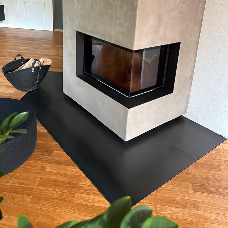 Funkenschutzplatte aus Schwarzstahl mit passgenauem Ausschnitt, montiert vor einem Kamin zum Schutz des Bodens.