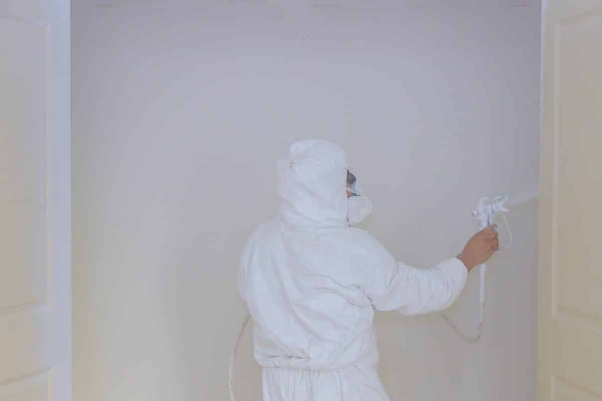 Person im weißen Schutzanzug lackiert Wand mit Sprühgerät.