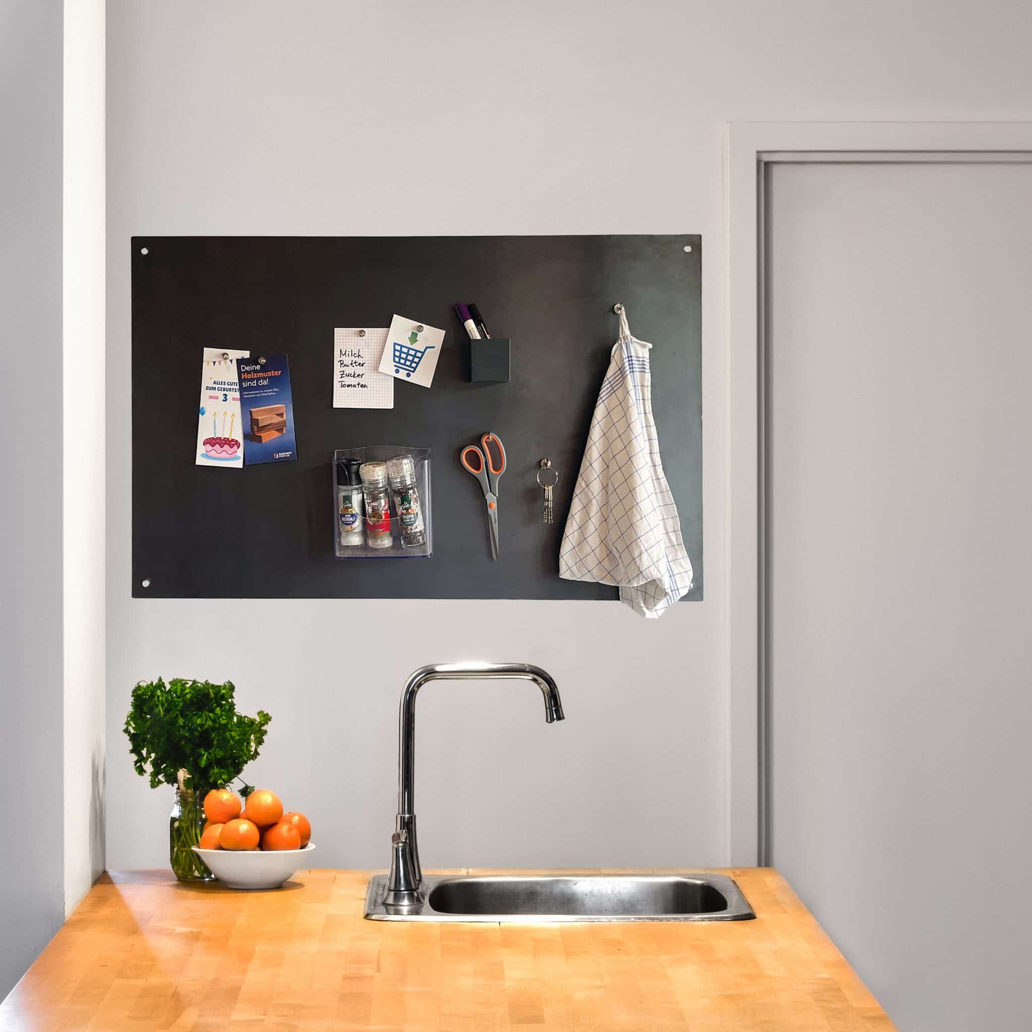 Eine schwarze Magnettafel hängt über einer hellen Küchenarbeitsplatte, auf der Pflanzen und Küchenutensilien ordentlich arrangiert sind.