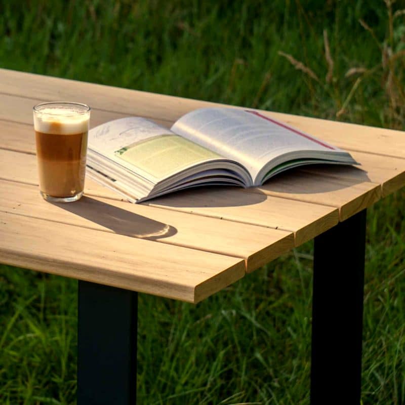 Holztisch im Außenbereich mit einem Buch und einem Glas Kaffee in der Sonne.