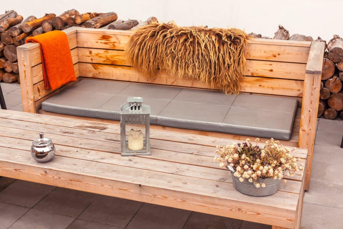 Holzbank mit grauem Polster sowie Holztisch mit Deko auf Terrasse.